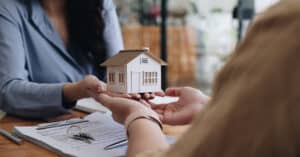 A female real estate agent or property professional hands a model home figure over to a customer with signed papers on the table below